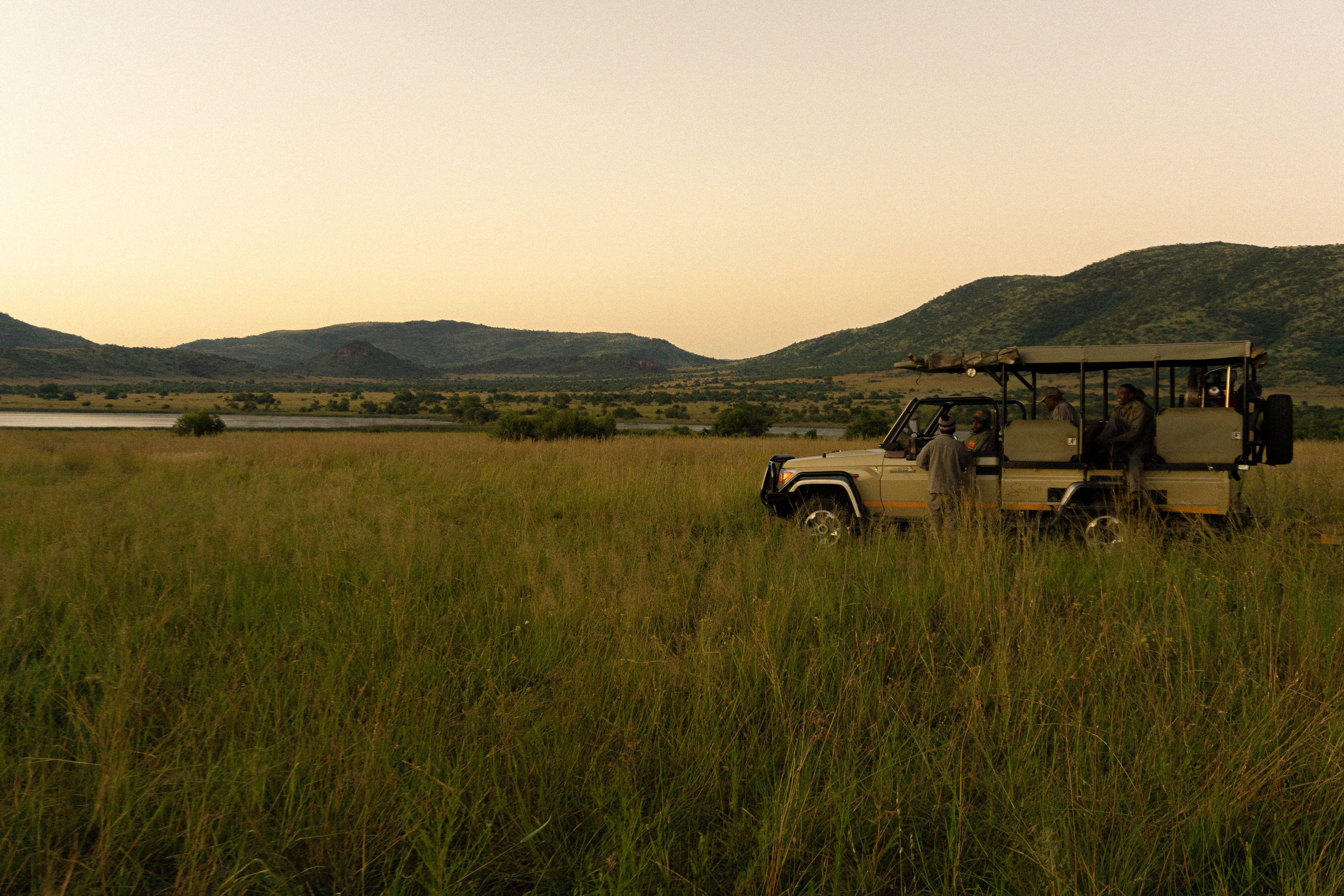 Safari guests and guide beside their game drive vehicle at dusk, mountains in the background